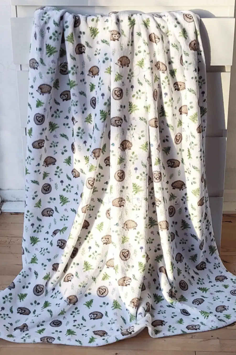 A soft, white fleece blanket featuring a charming pattern of brown hedgehogs and green ferns.