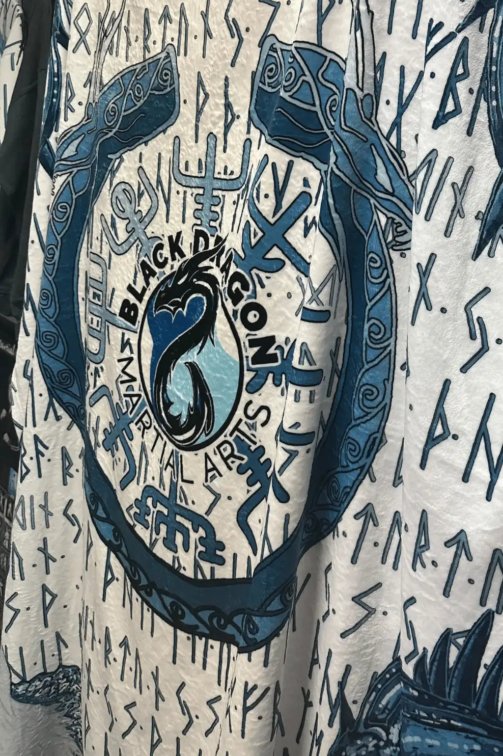 White fabric garment featuring a bold blue circular emblem with a stylized dragon and the text ’black dragon martial arts,’ surrounded by intricate rune-like patterns.